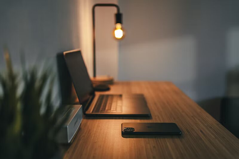 Un angolo di lavoro accogliente e organizzato con un laptop elegante, uno smartphone e una lampada da scrivania calda. La scrivania in legno e l'illuminazione soffusa creano un'atmosfera concentrata e invitante, perfetta per il lavoro remoto o la creatività notturna.