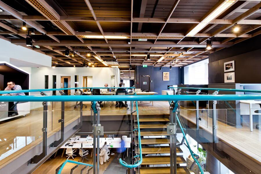 Ufficio open space con arredi moderni, soffitto in legno a griglia, scala interna in vetro e metallo, e postazioni di lavoro condivise